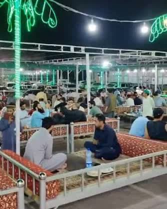 The outdoor majlis at night, charpai tables full of guests under strings of lights